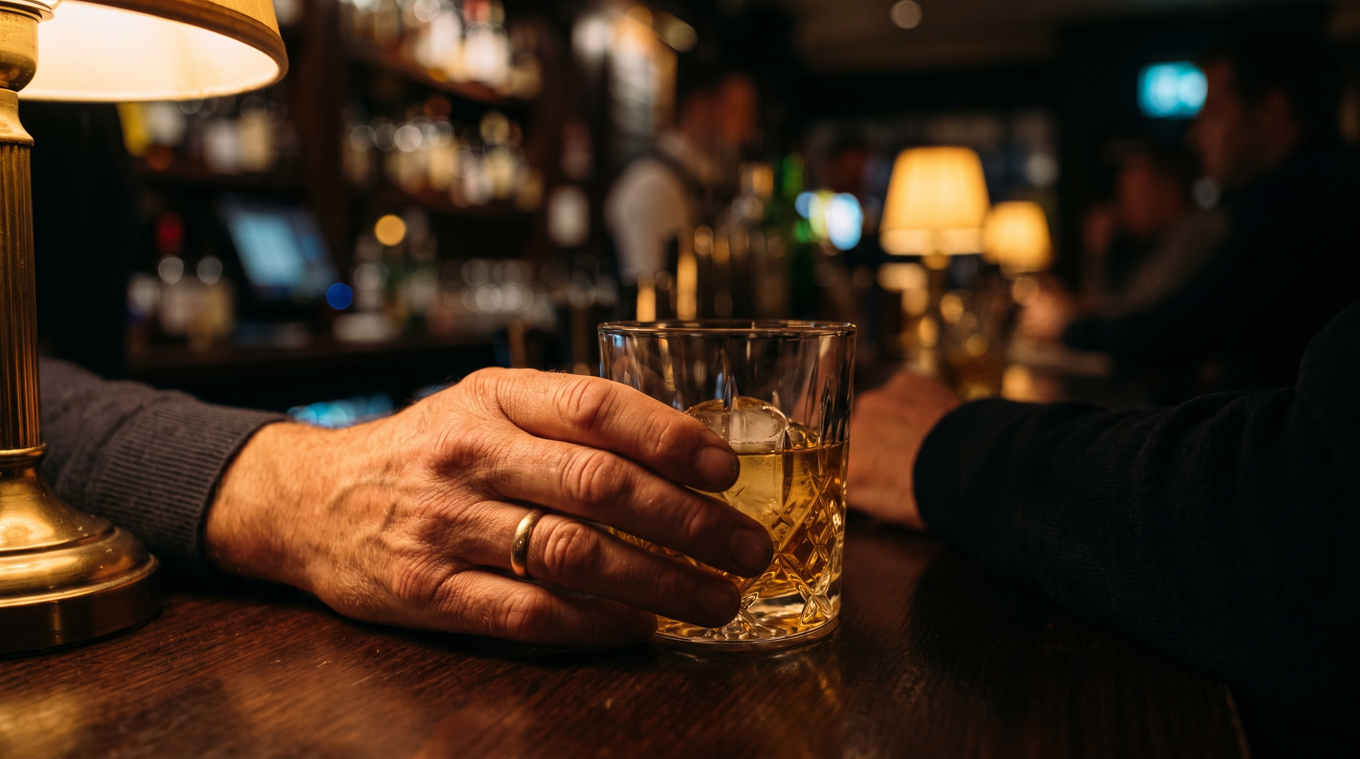 Main d'homme tenant un verre de whisky ambré sur un comptoir de bar en bois, lumière tamisée